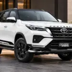 White 2026 Toyota Fortuner Leader Edition with black alloy wheels, a black roof and bold front grille outside a modern building.